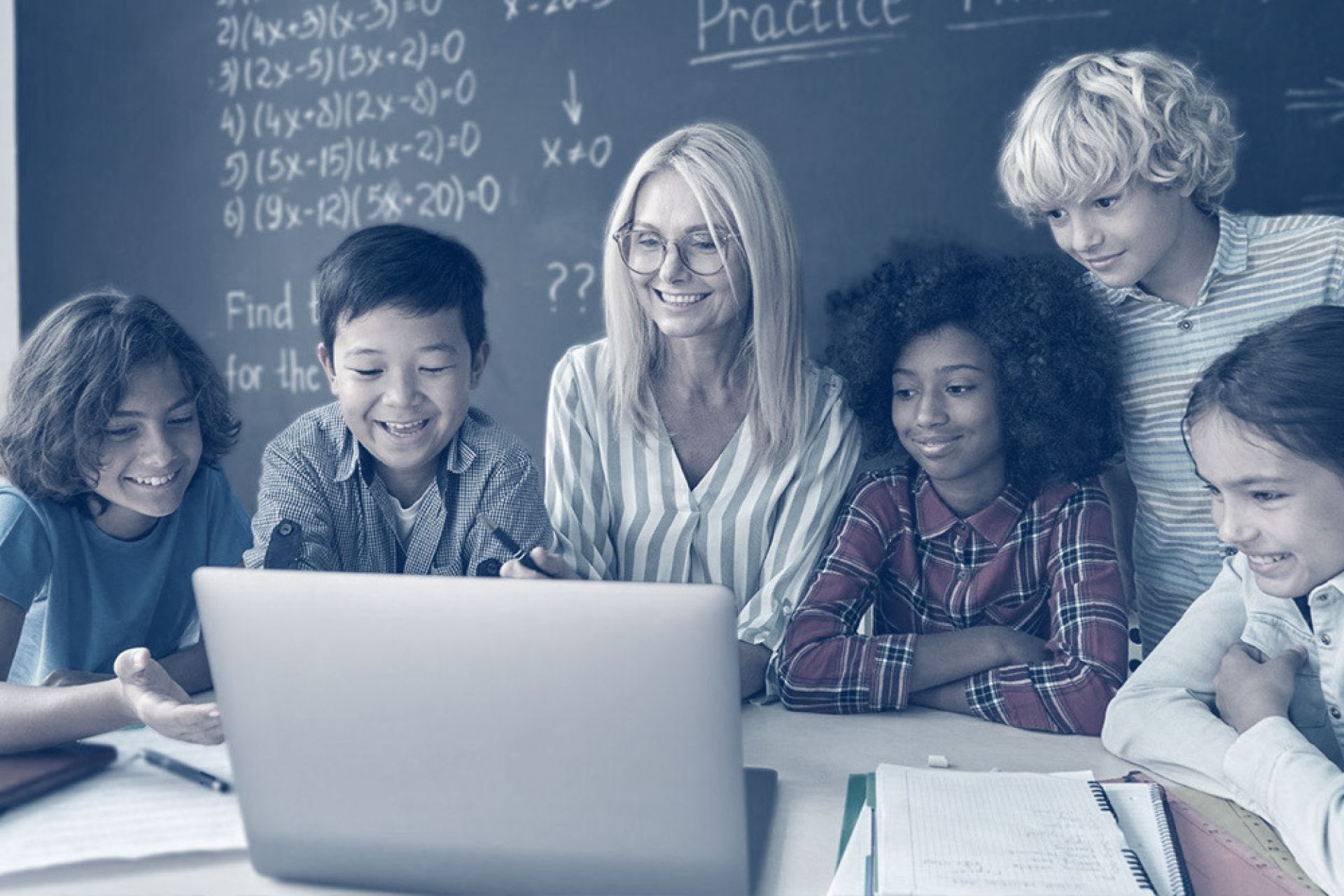 Teacher seated at desk with open laptop surrounded by older elementary students looking at her screen enthusiastically.